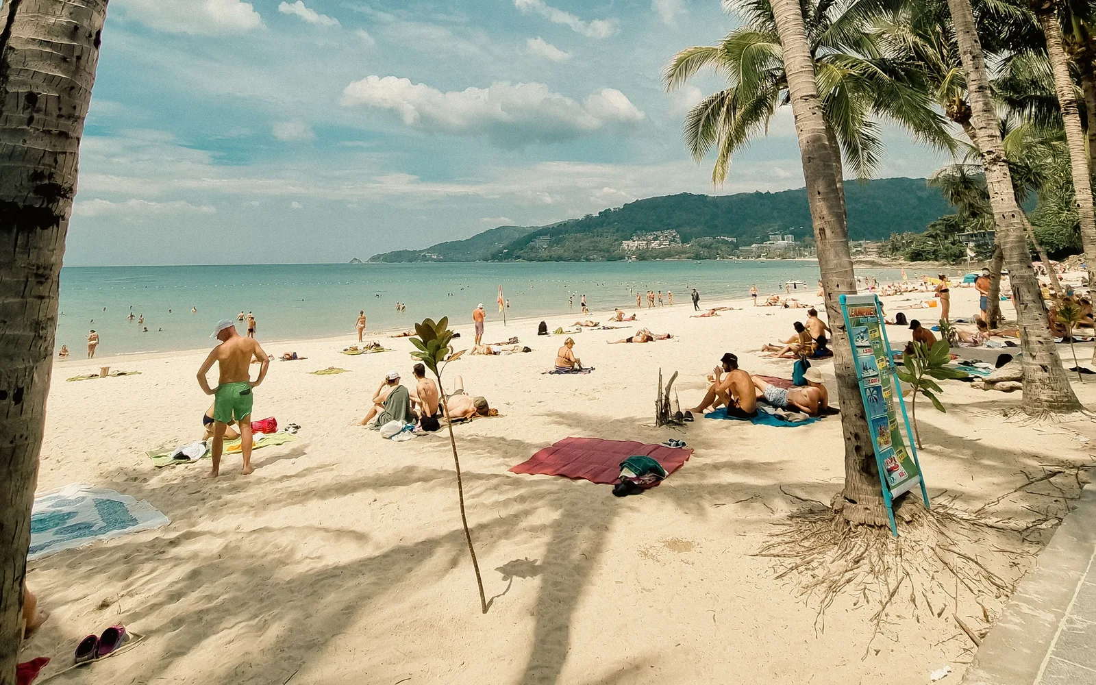 Sunny day at Patong Beach, Phuket, Thailand, featuring palm trees, sand, and relaxing tourists.